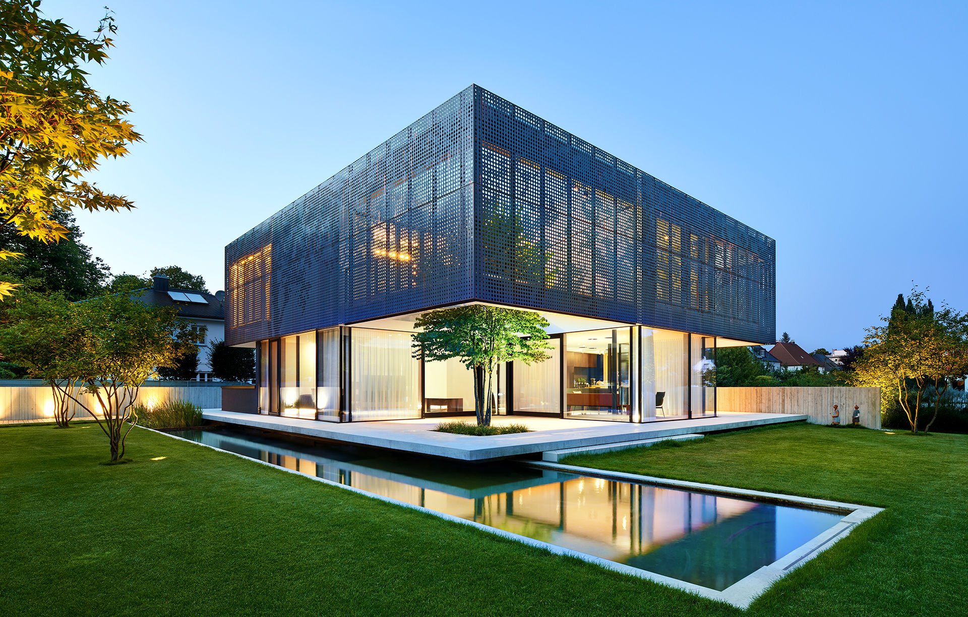 Modernes, kubisches Wohnhaus mit Glasfassade und Metallverkleidung, beleuchtet bei Dämmerung mit Wasserbecken im Garten