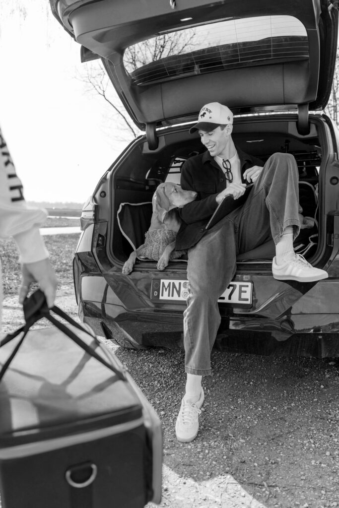 Mann sitzt mit Hund im Kofferraum eines Autos und lacht